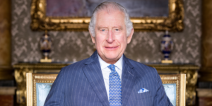 A recent photograph of King Charles III. He wears a blue suit, and smiles directly at the camera.